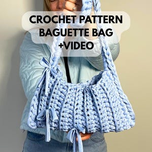 Op de afbeelding: Een lichtblauwe gehaakte baguette tas met een gevlochten band en strikaccenten. De tas is gemaakt van dik, getextureerd garen. De tekst "CROCHET PATTERN BAGUETTE BAG +VIDEO" is zichtbaar.