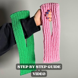 May include: Two knitted arm warmers, one bright green and one pink, are held up against a gray background. The arm warmers have a ribbed texture and a thumb hole. Text at the bottom reads "STEP BY STEP GUIDE + VIDEO".