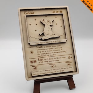 May include: A wooden Pokemon trading card featuring Cubone, a Ground-type Pokemon. The card has a brown background with a white border and features a picture of Cubone in the center. The card also includes the Pokemon's name, type, HP, attacks, weakness, resistance, and retreat cost. The text on the card reads: "Cubone, Lonely Pokemon. Length: 4" Weight: 14 lbs. Snivel: If the Defending Pokemon attacks Cubone during your opponent's next turn, any damage done by the attack is reduced by 20 (after applying Weakness and Resistance). (Benching either Pokemon ends this effect). Rage: Does 10 damage plus 10 more damage for each damage counter on Cubone. Weakness: Grass. Resistance: Electric -30. Retreat Cost: 1 Star. Because it never removes its skull helmet, no one has ever seen this Pokemon's real face. LV. 13 #104."