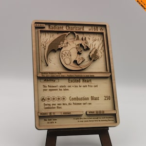 May include: A wooden Radiant Charizard Pokémon card replica, featuring a detailed laser-cut design. The card displays the character's name, HP, abilities, and attack details. It is mounted on a small wooden easel, showcasing the card's artwork.