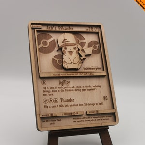 Op de afbeelding: Houten replica van een Ash's Pikachu Pokémon-kaart, met een 3D Pikachu-ontwerp met een hoed. De kaart wordt weergegeven op een kleine houten ezel. De tekst bevat "I Choose You" en kaartdetails.
