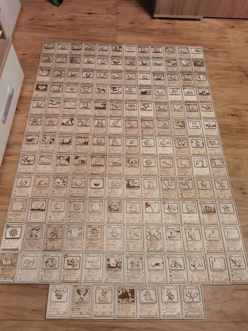 May include: A collection of 100 wooden Pokemon cards featuring various Pokemon characters. The cards are arranged in a grid pattern on a wooden floor.