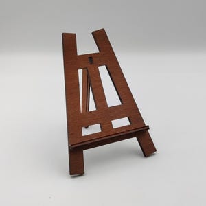 May include: A small wooden easel with a brown finish. The easel has a rectangular base and a triangular top with a crossbar for holding a canvas or picture.