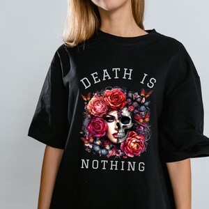 May include: Black t-shirt featuring a graphic design with a skull, roses, and butterflies. The text "DEATH IS NOTHING" is printed above and below the graphic. The design incorporates red, pink, and white colors.