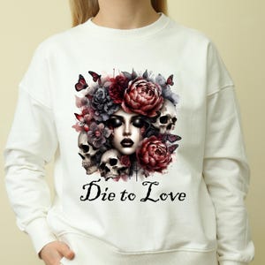 May include: White sweatshirt featuring a watercolor design of a woman's face surrounded by skulls, flowers, and butterflies. The phrase "Die to Love" is printed in black script below the image. The design has a gothic and romantic theme.