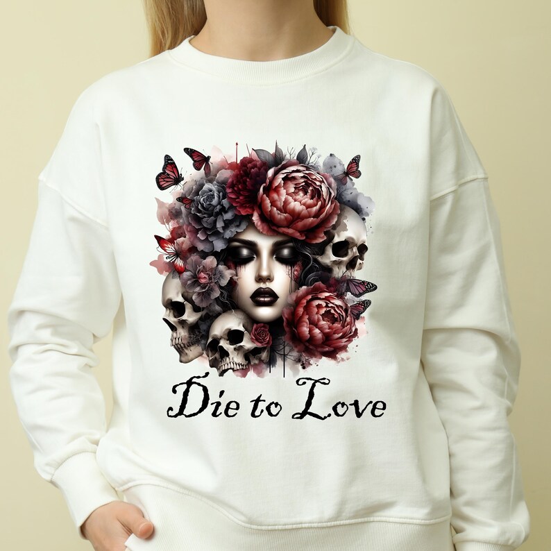 May include: White sweatshirt featuring a watercolor design of a woman's face surrounded by skulls, flowers, and butterflies. The phrase "Die to Love" is printed in black script below the image. The design has a gothic and romantic theme.