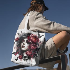 May include: White tote bag featuring a watercolor design of a woman's face, surrounded by red peonies, butterflies, and a skull. The bag has a light gray handle and is made of a durable fabric. The design has a gothic and romantic theme.