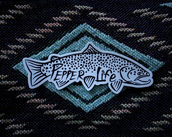 May include: A black and white sticker of a trout fish with the text "Pepper Life" on it. The sticker is on a blue and brown patterned fabric.