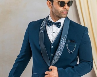 May include: A teal blue suit with a black satin lapel and a white shirt with a black bow tie. The suit has a decorative chain and a patterned design on the lapel. The man is wearing black loafers.