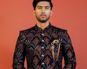 Könnte beinhalten: Ein Mann trägt eine lange, schwarze, gemusterte Kurta mit goldenen Knöpfen und einer goldenen Kette. Er trägt auch beige Hosen und braune Schuhe. Der Hintergrund ist einfarbig rot.
