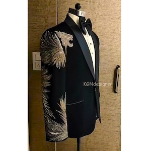 Könnte beinhalten: Ein schwarzer Smoking-Sakko mit Satin-Revers und einem gold- und silberfarbenen Stickmuster auf dem linken Ärmel. Das Muster zeigt einen stilisierten Tierkopf und Federn.