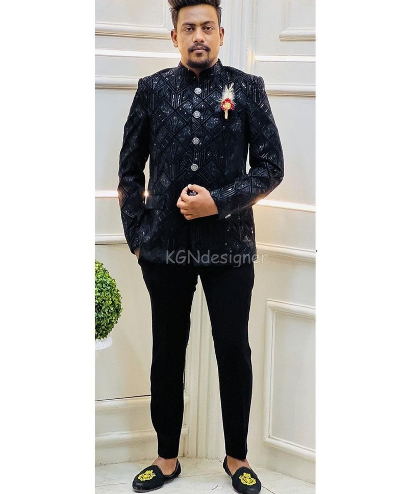 K&ouml;nnte beinhalten: Ein schwarzer Blazer mit einem Paillettenmuster und einem Stehkragen. Der Blazer hat einen Knopfverschluss und wird &uuml;ber einer schwarzen Hose getragen.