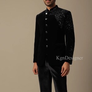 May include: A black velvet suit jacket with a mandarin collar and a silver embroidered design on the right side. The jacket has a single button closure and a pocket on the left side. The suit is paired with black velvet pants.