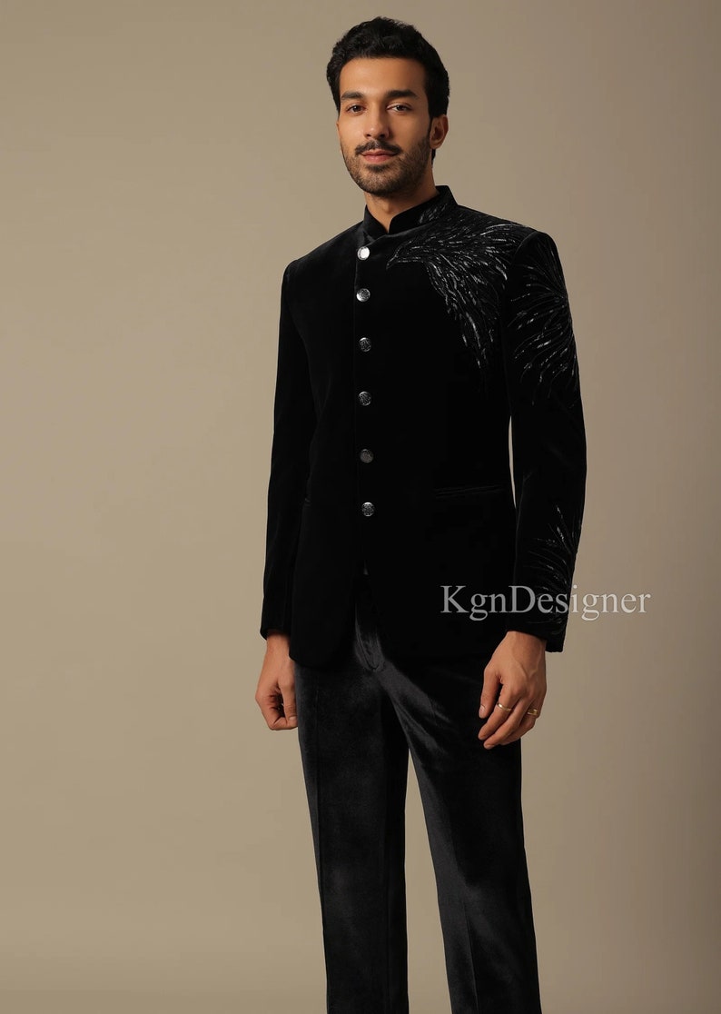 May include: A black velvet suit jacket with a mandarin collar and a silver embroidered design on the right side. The jacket has a single button closure and a pocket on the left side. The suit is paired with black velvet pants.