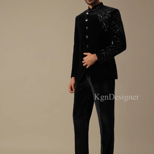 May include: A black velvet suit with a mandarin collar and a silver embroidered design on the jacket. The suit is worn by a person standing on a light brown background.