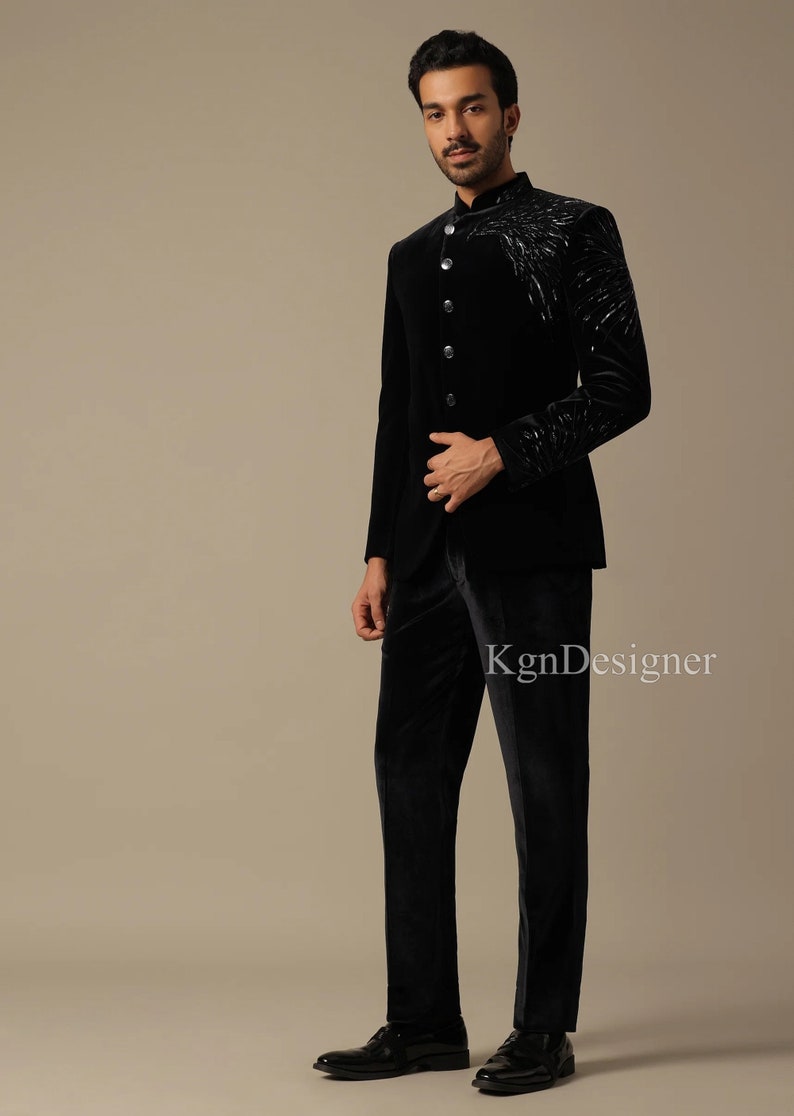 May include: A black velvet suit with a mandarin collar and a silver embroidered design on the jacket. The suit is worn by a person standing on a light brown background.