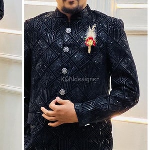 K&ouml;nnte beinhalten: Eine schwarze Samtjacke mit geometrischem Muster und silbernen Kn&ouml;pfen. Die Jacke hat einen Stehkragen und einen einzigen Knopfverschluss. Eine kleine Blumennadel befindet sich am Revers.