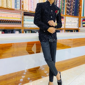 K&ouml;nnte beinhalten: Ein schwarzer Blazer mit einem Paillettenmuster und einer kleinen roten Blume am Revers. Der Blazer wird &uuml;ber einem schwarzen Hemd mit Kragen und einer schwarzen Hose getragen. Die Person tr&auml;gt schwarze Schuhe.