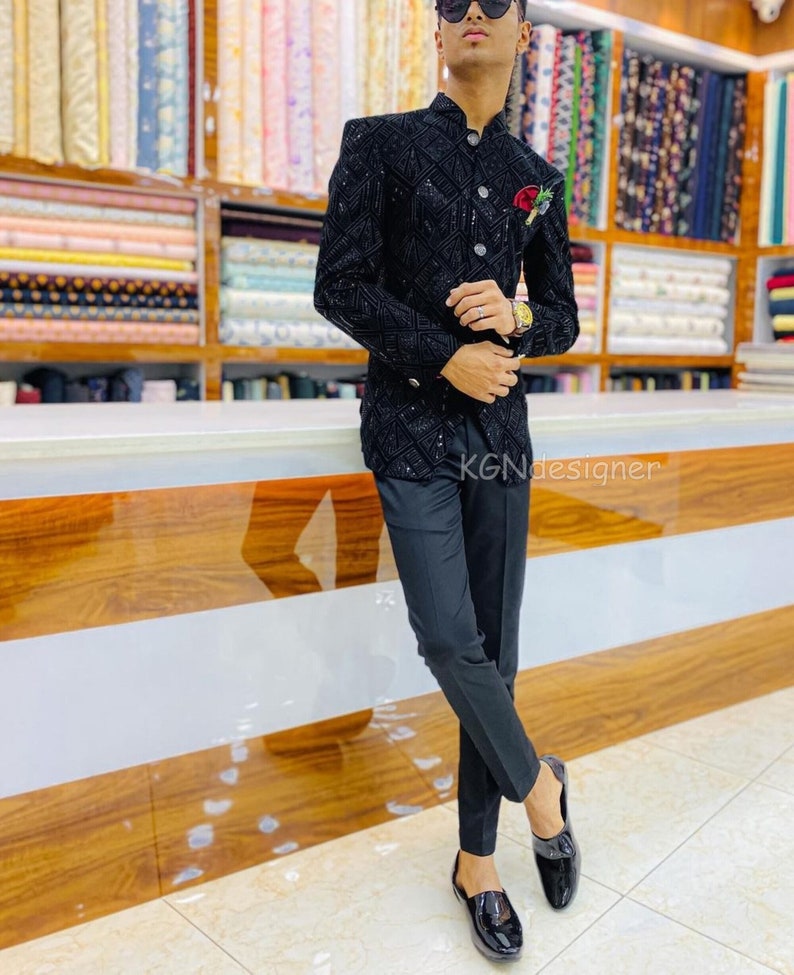 K&ouml;nnte beinhalten: Ein schwarzer Blazer mit einem Paillettenmuster und einer kleinen roten Blume am Revers. Der Blazer wird &uuml;ber einem schwarzen Hemd mit Kragen und einer schwarzen Hose getragen. Die Person tr&auml;gt schwarze Schuhe.