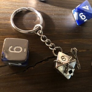 May include: A silver keychain with a silver dice charm and a silver key charm. The dice charm has the number 8 on it. The keychain is attached to a silver ring.