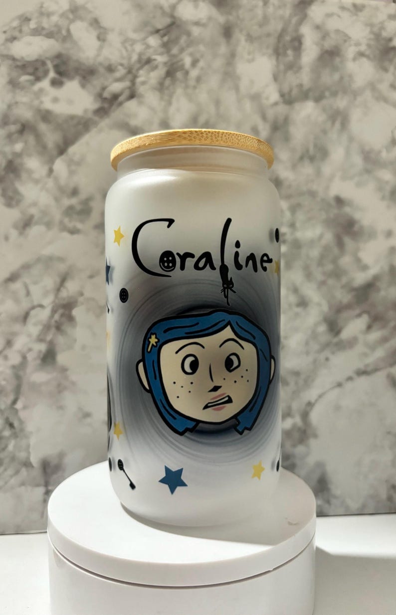 Pu&ograve; includere: Un barattolo di vetro bianco con un coperchio in bamb&ugrave;, con un design dipinto a mano di Coraline dal film Coraline. Il design include una spirale in bianco e nero, stelle e un bottone.