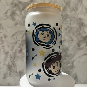 Pu&ograve; includere: Vaso in ceramica bianca con coperchio in legno, decorato con un motivo in bianco e nero che rappresenta i personaggi del film "Coraline". Il motivo comprende stelle, chiavi e bottoni.