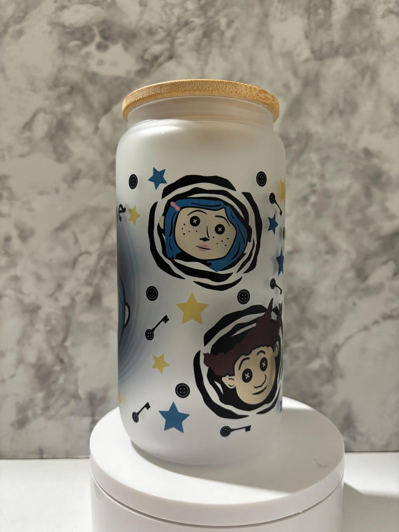 Pu&ograve; includere: Vaso in ceramica bianca con coperchio in legno, decorato con un motivo in bianco e nero che rappresenta i personaggi del film "Coraline". Il motivo comprende stelle, chiavi e bottoni.