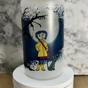 Pu&ograve; includere: Un barattolo di vetro smerigliato con un coperchio in bamb&ugrave; che presenta un'illustrazione di cartone animato di una ragazza con un impermeabile giallo e un gatto nero in piedi di fronte a una luna piena. Lo sfondo &egrave; blu scuro con schizzi di vernice bianca.