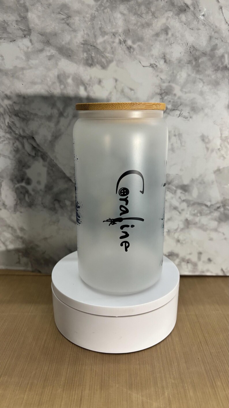 Pu&ograve; includere: Un barattolo di vetro smerigliato con un coperchio in bamb&ugrave;. Il barattolo ha un design in bianco e nero e il testo "Coraline" sul lato.