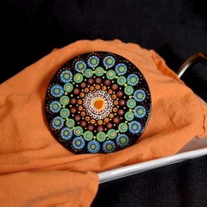 May include: A black, round coaster with a colorful mandala design. The mandala features concentric circles of blue, green, orange, and yellow dots. The coaster is resting on an orange cloth.