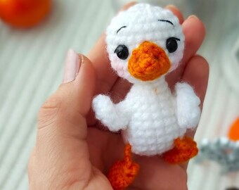 May include: A small white crocheted duck with orange feet and beak. The duck is being held in a person's hand.