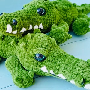 May include: Two green crocheted alligator plushies with large black eyes and white teeth. The plushies are lying on a light blue surface.