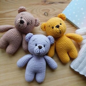 Op de afbeelding: Drie gehaakte teddyberen in verschillende kleuren: lichtbruin, geel en lavendel. De beren zitten allemaal op een houten oppervlak.