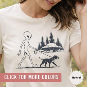 May include: A natural-colored t-shirt featuring a black and white graphic of an alien walking a dog on a leash. The design includes a UFO and trees in the background. The text "CLICK FOR MORE COLORS" is displayed on the bottom.