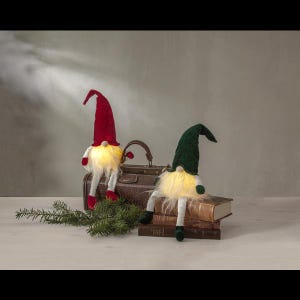 Könnte beinhalten: Zwei dekorative Zwerge mit beleuchteten Bäuchen und langen weißen Bärten. Einer trägt einen roten Hut, der andere einen grünen Hut. Sie posieren mit einem Vintage-Koffer und Büchern, mit einem Zweig Grün.