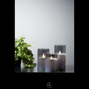 Könnte beinhalten: Drei graue Glas-Kerzenhalter mit flackernden LED-Kerzen. Die Kerzen sind in verschiedenen Größen und auf einer schwarzen Oberfläche angeordnet.