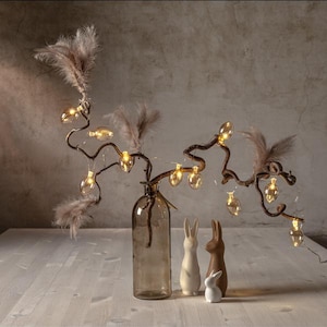 Könnte beinhalten: Ein brauner Ast mit kleinen, klaren Glasbirnen, die mit Lichtern besetzt sind, in einer klaren Glasvase. Zwei weiße Keramikhasen und ein brauner Keramikhase stehen vor der Vase.