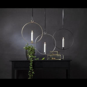 Könnte beinhalten: Drei hängende Kerzenhalter mit runden Rahmen. Die Rahmen sind aus Metall und in Gold, Silber und Schwarz gehalten. Jeder Rahmen hält eine weiße Kerze.