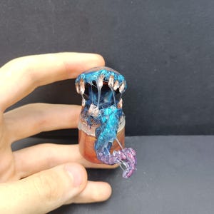 May include: A small, cylindrical container topped with a sculpted creature featuring a wide, open mouth with sharp, light-colored teeth. The creature has a glossy blue and purple finish with translucent, stringy, web-like resin details extending downward.