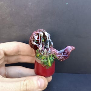 May include: A small, cylindrical red container topped with a sculpted, organic-looking mass in shades of deep purple, green, and red. The sculpture features translucent, stringy white webbing connecting its textured, fleshy-looking protrusions.