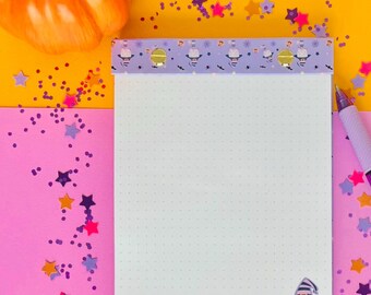 May include: A purple and white notepad with a witch and stars design. The notepad is on a pink and yellow background with purple and orange glitter.