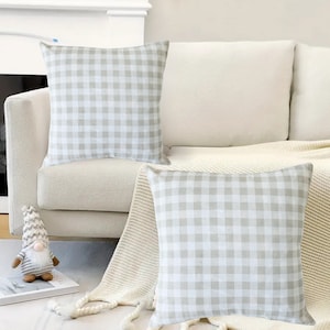 May include: Two beige and white gingham throw pillows on a white couch with a white throw blanket.