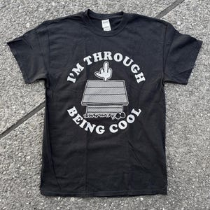 Op de afbeelding: Zwart T-shirt met een witte afbeelding van een hondenhok met een hand die uit het dak steekt. De tekst "I'm Through Being Cool" is rond de afbeelding gedrukt.