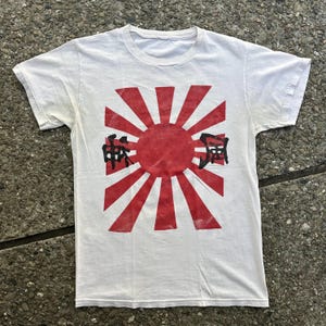 May include: White t-shirt featuring a red and white rising sun graphic with the Japanese characters "神風" printed in black.