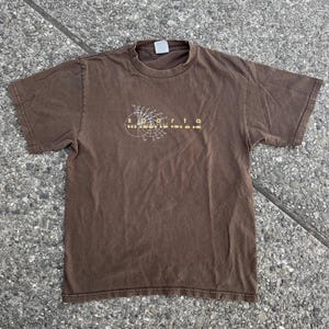 Puede incluir: Una camiseta marrón con un diseño gráfico de un círculo con líneas y la palabra "s.o.a.r.t.f.a." impresa en amarillo.