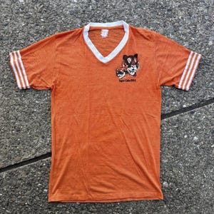 Könnte beinhalten: Orangefarbenes Vintage-T-Shirt mit weißem V-Ausschnitt und weißen Streifen auf den Ärmeln. Das Shirt zeigt eine Grafik von zwei Tigergesichtern und den Text "Tiger Cubs BSA" in Schwarz.