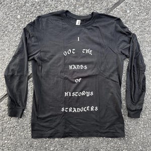 Puede incluir: Camiseta negra de manga larga con texto blanco que dice "I got the hands of history's stranglers".