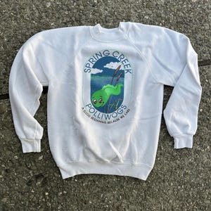Puede incluir: Sudadera blanca con un logotipo verde y azul que dice "Spring Creek Polliwogs" y "A good beginning because we care".