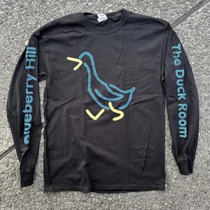 Puede incluir: Camiseta negra de manga larga con un contorno amarillo y azul de un pato en la parte delantera. La manga izquierda tiene el texto "Blueberry Hill" impreso verticalmente y la manga derecha tiene el texto "The Duck Room" impreso verticalmente.