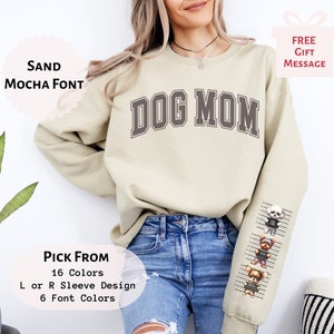 May include: A beige sweatshirt with the text "DOG MOM" in a dark gray font. The sleeves have a line-up of cartoon dogs in a jail cell design. The text "PICK FROM 16 Colors L or R Sleeve Design 6 Font Colors" is on the bottom left of the image.